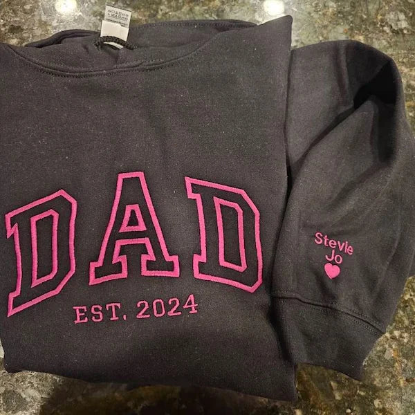 Black hoodie with pink 'DAD EST. 2024' text on a stone surface