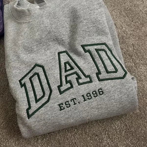 Gray sweatshirt with 'DAD EST. 1996' text on a beige carpet