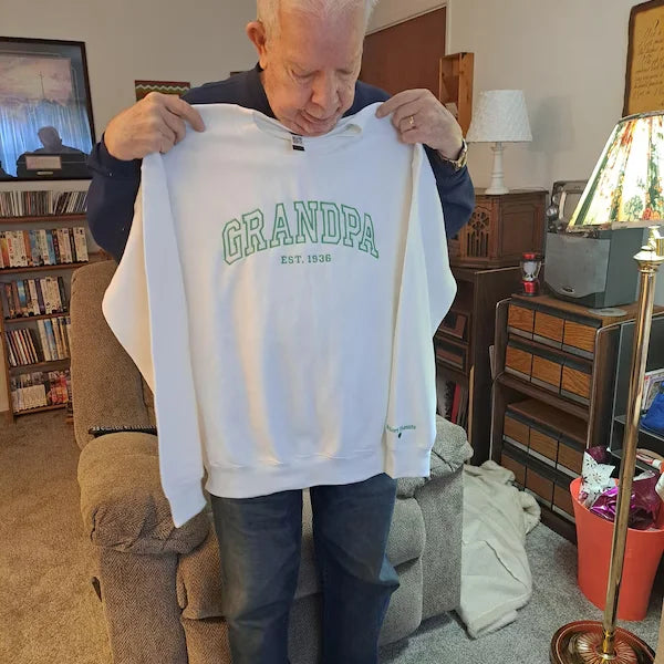 Man holding a white sweatshirt with 'GRANDPA' text in a living room setting.