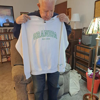 Man holding a white sweatshirt with 'GRANDPA' text in a living room setting.