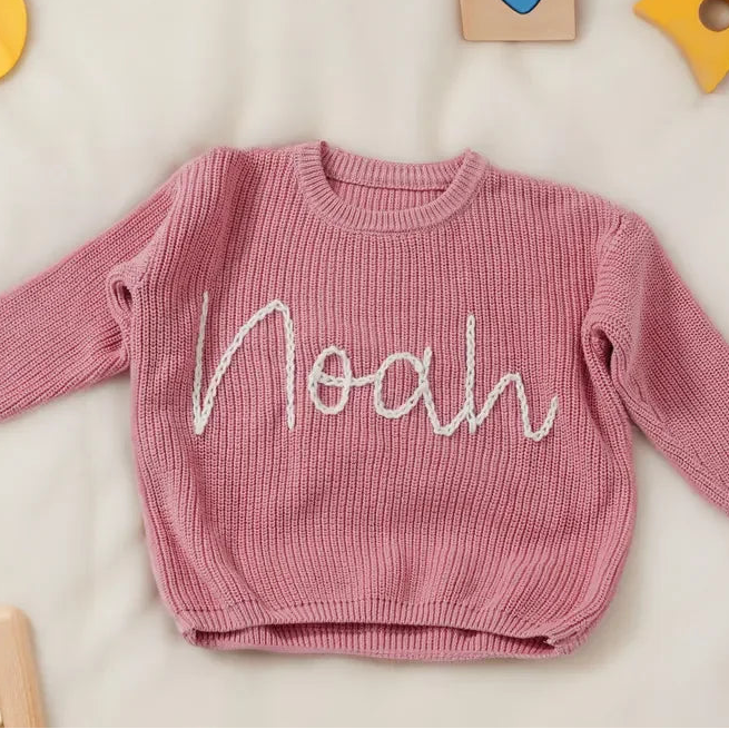 Pink sweater with 'Noah' embroidery on a light background with toys around