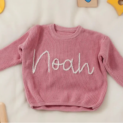 Pink sweater with 'Noah' embroidery on a light background with toys around