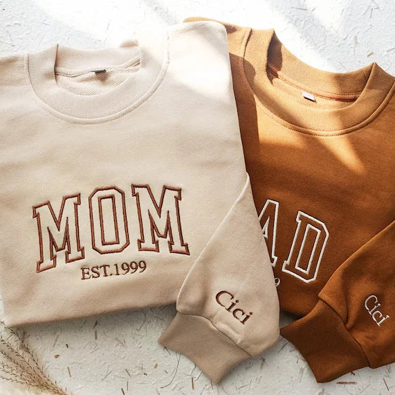 Two folded sweatshirts, one beige with 'MOM EST.1999' text and the other brown with 'DAD' text, on a textured white surface.