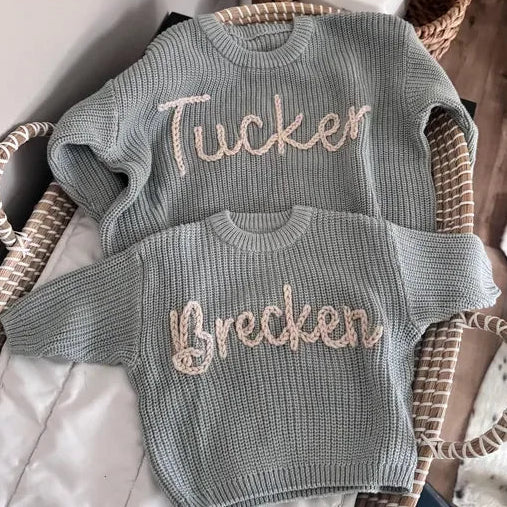 Two gray sweaters with embroidered names on a wicker chair.