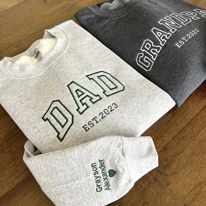 Two gray sweatshirts with 'DAD EST. 2023' and 'GRANDPA EST. 2023' text on a wooden surface.