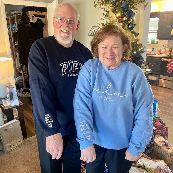Two people wearing sweatshirts with text standing in a home setting with a Christmas tree.