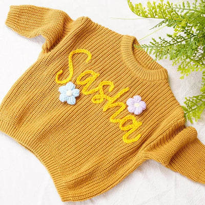 Yellow knitted sweater with 'Sasha' and floral designs on a white background with greenery.